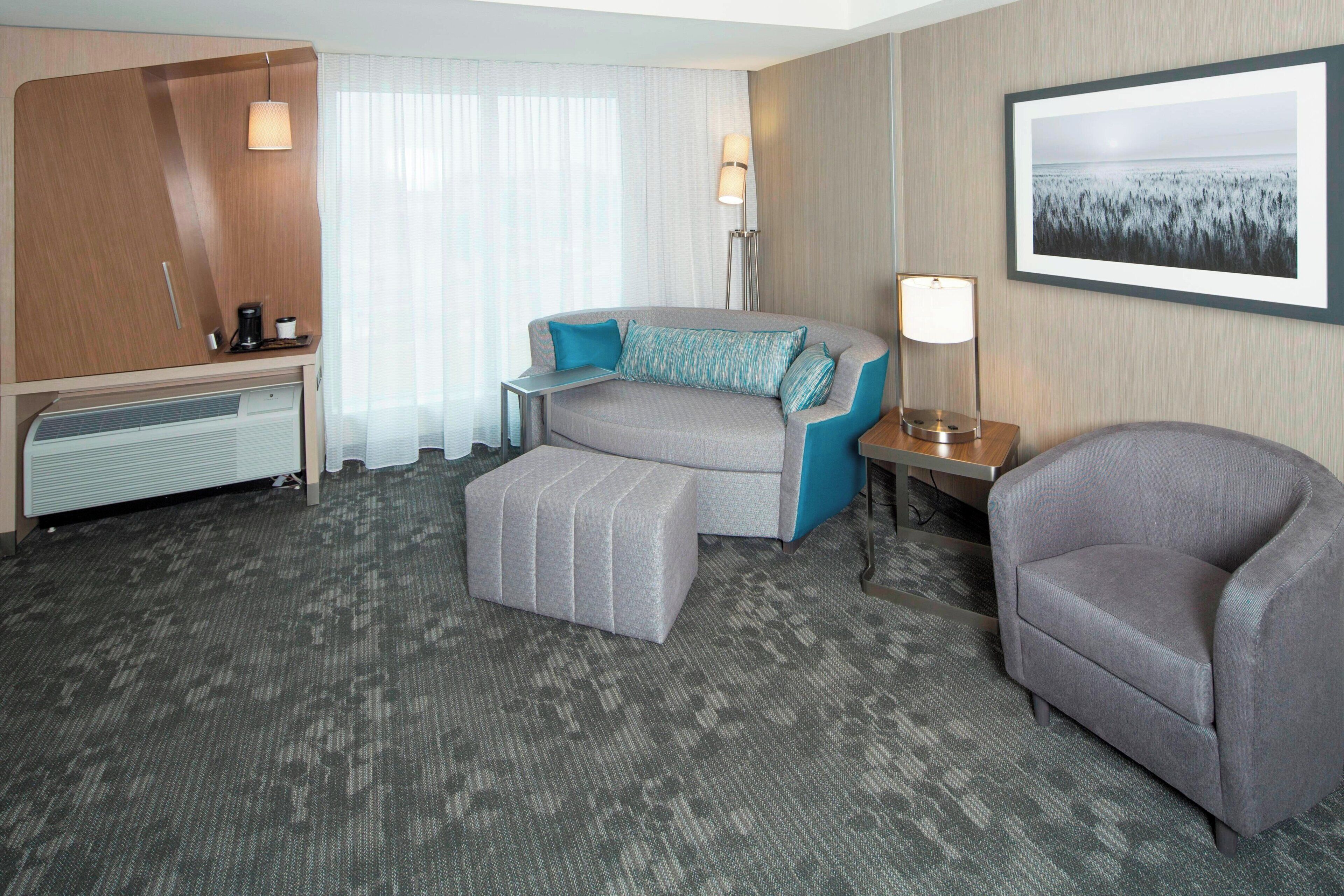 A hotel room with a sofa, ottoman, armchair, bedside table, lamp, and a framed picture on the wall.