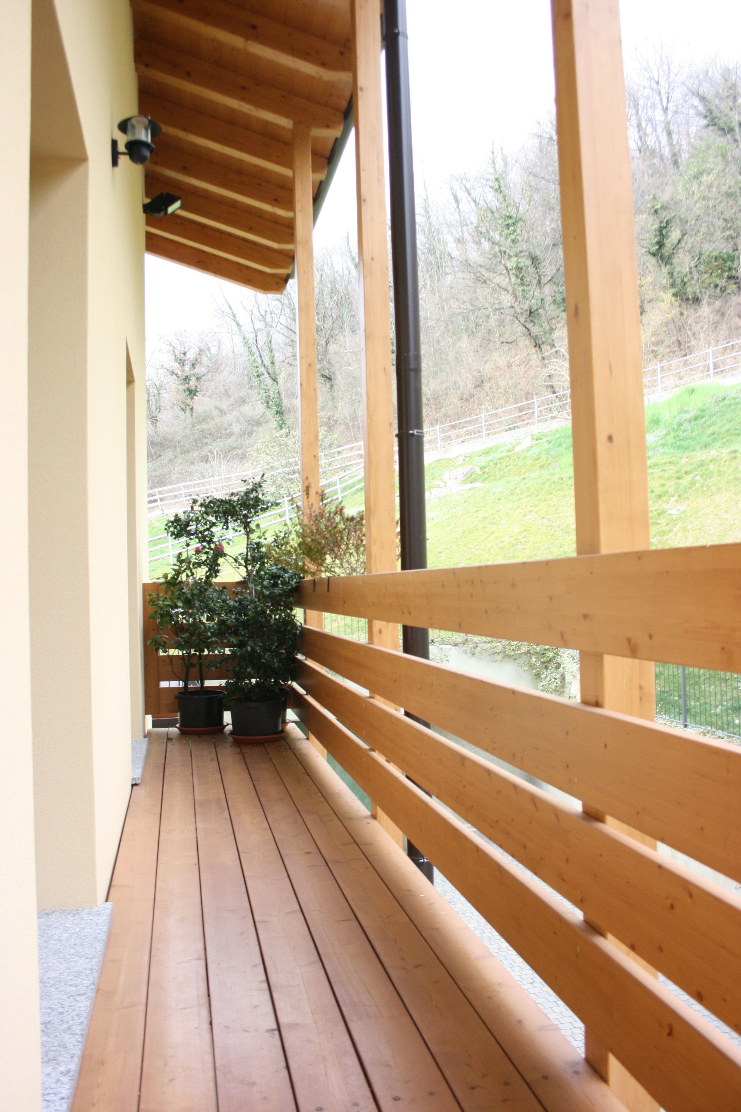 Una terraza de madera con un banco, plantas en macetas y vistas a un área verde más allá de una valla.