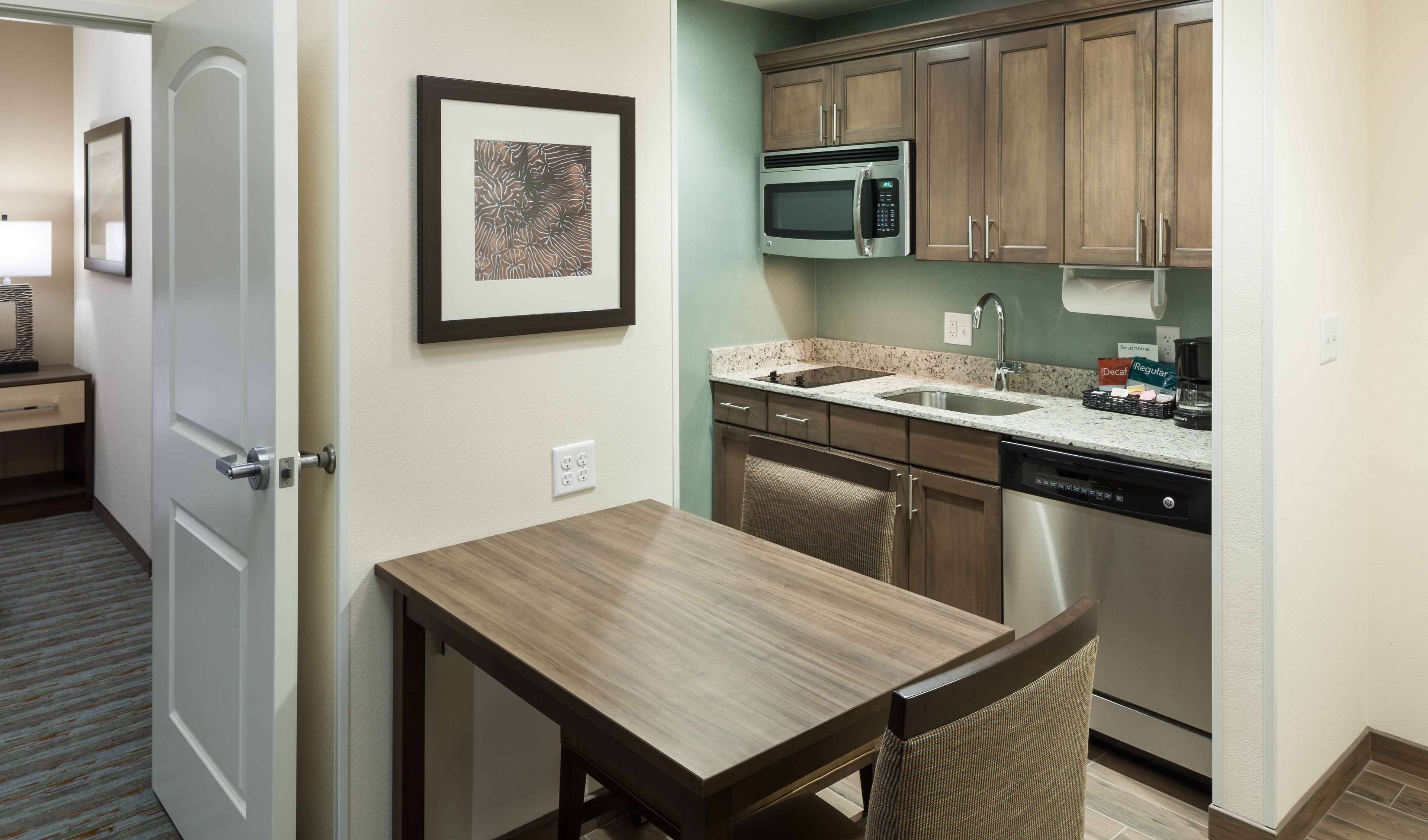 A compact kitchen with a microwave, sink, and dishwasher. There's a small table and a chair, and a framed artwork on the wall.