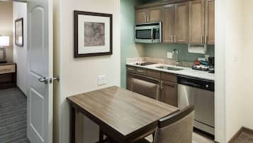 A compact kitchen with a microwave, sink, and dishwasher. There's a small table and a chair, and a framed artwork on the wall.