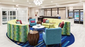 A modern lobby with a seating area featuring a yellow sofa, teal armchairs, and a coffee table. There are vending machines and a reception desk in the background. - Homewood Suites by Hilton Cape Canaveral-Cocoa Beach (Cape Canaveral)
