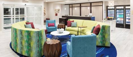 A modern lobby with a seating area featuring a yellow sofa, teal armchairs, and a coffee table. There are vending machines and a reception desk in the background.