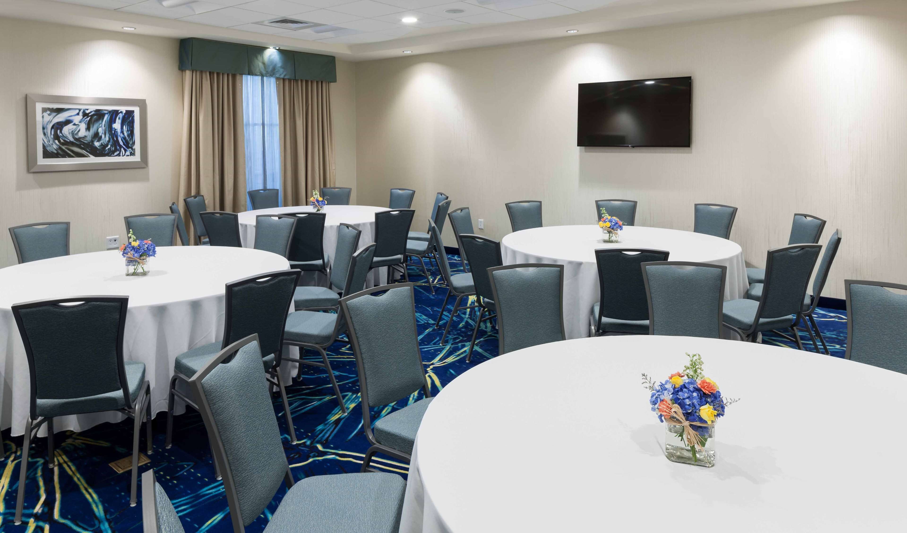 A conference room with multiple round tables, grey chairs, and a flat-screen TV on the wall.