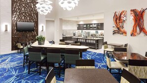 A hotel dining area with a central round table, surrounded by chairs with striped cushions. A flat-screen TV is mounted on the wall. The room features a blue patterned carpet and modern decor. - Homewood Suites by Hilton Cape Canaveral-Cocoa Beach (Cape Canaveral)