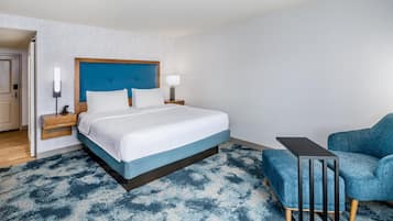 Quarto de hotel com cama, mesa de cabeceira, luminária e uma poltrona azul.
