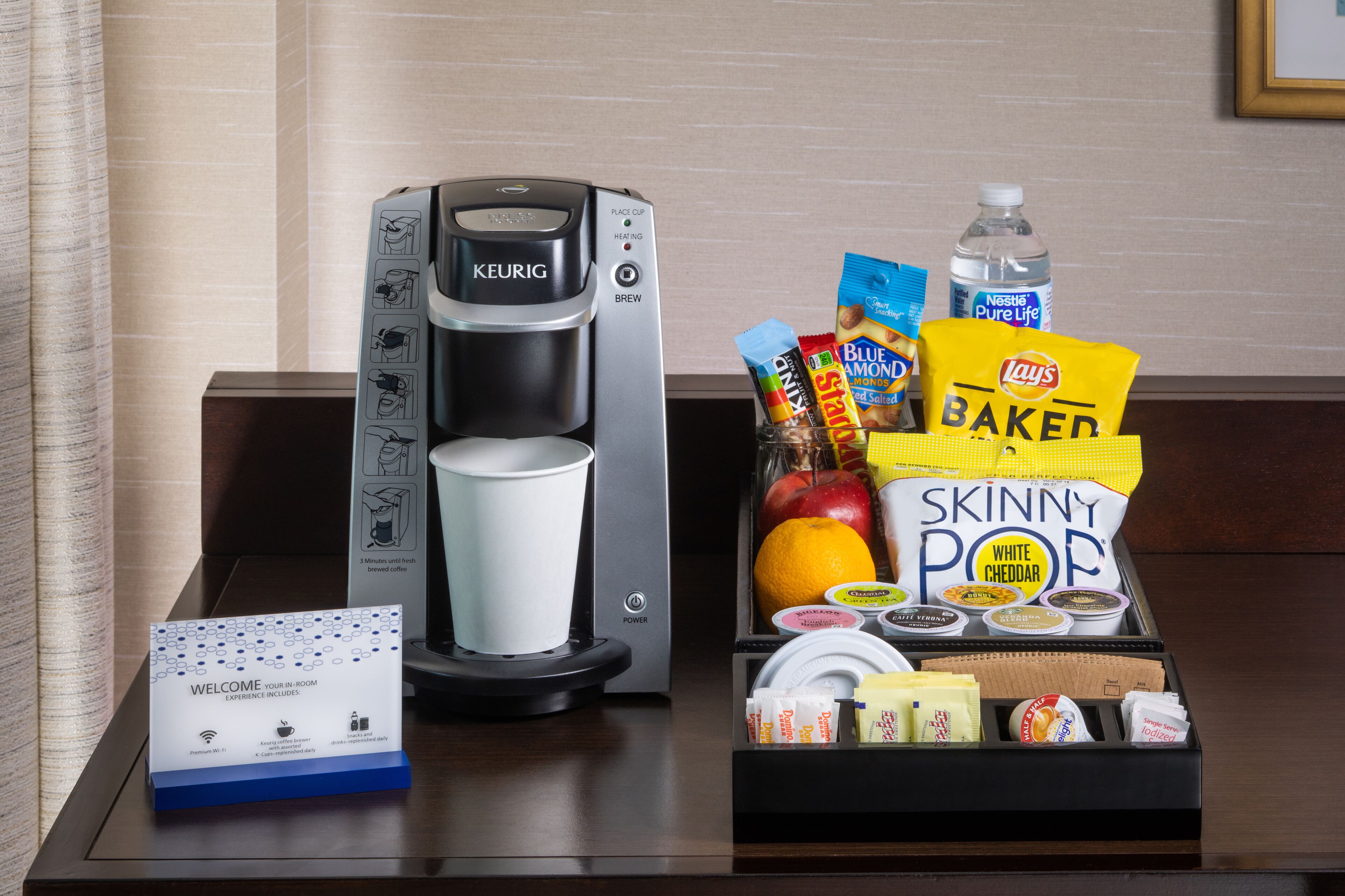 A coffee maker, a tray with snacks, and a welcome sign on a hotel table.