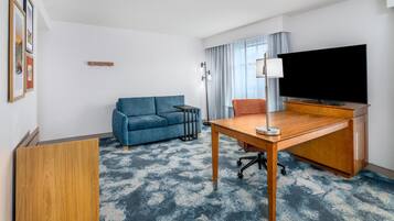 Une chambre d’hôtel avec un canapé bleu, un bureau en bois avec une télévision, une lampe et une grande fenêtre avec des rideaux.