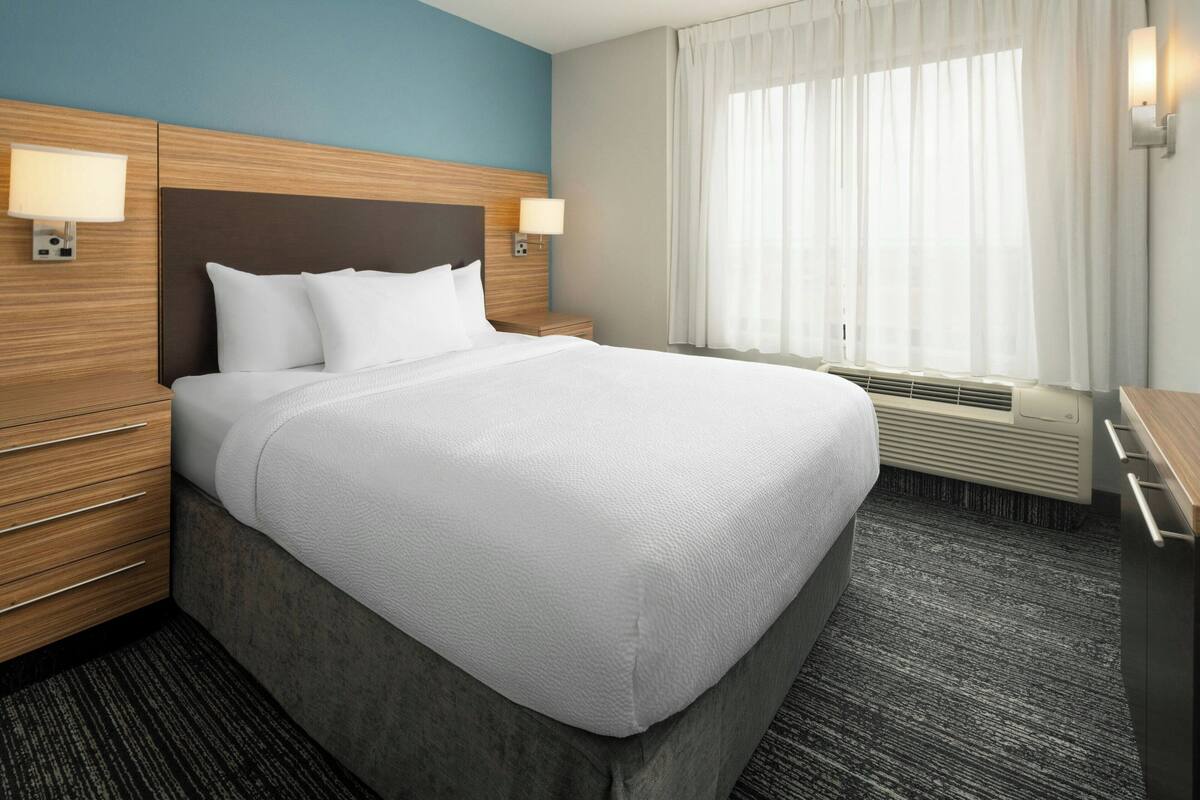 Habitación de hotel con una cama grande, dos lámparas de noche, una ventana con cortinas y un aire acondicionado de pared.