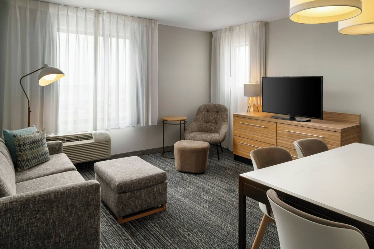 Una habitación de hotel con sofá, reposapiés, televisor, cómoda de madera y una lámpara.