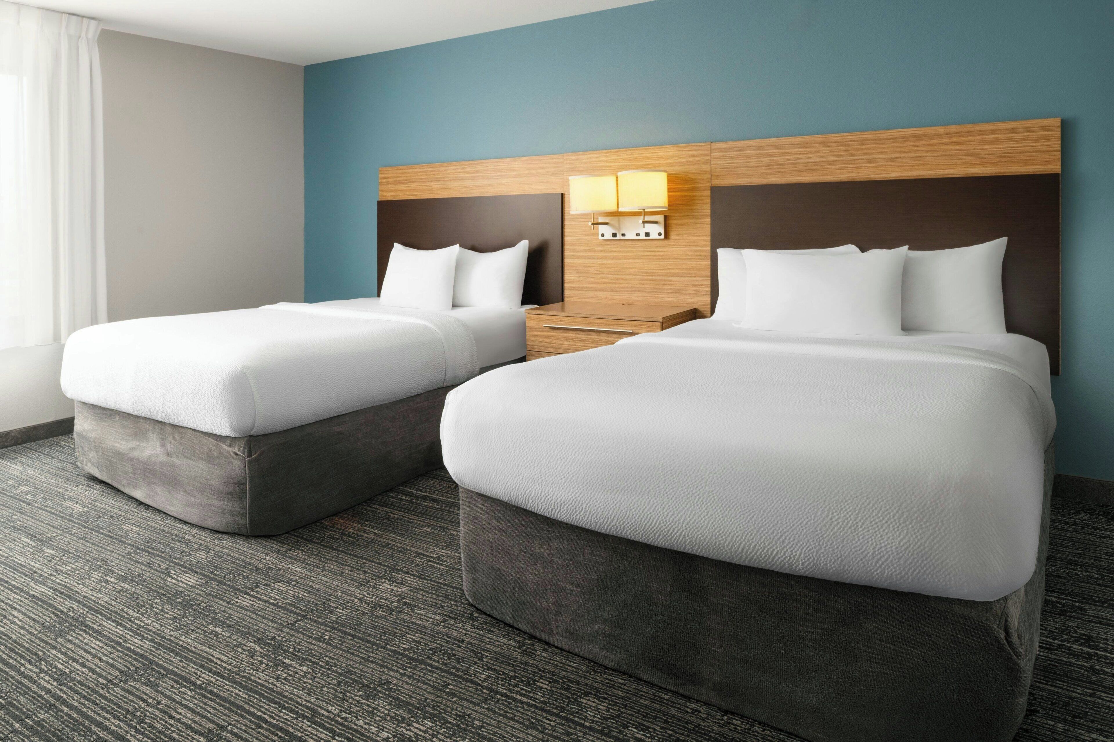 A hotel room with two beds, a wooden headboard, a nightstand with a lamp, and a carpeted floor.