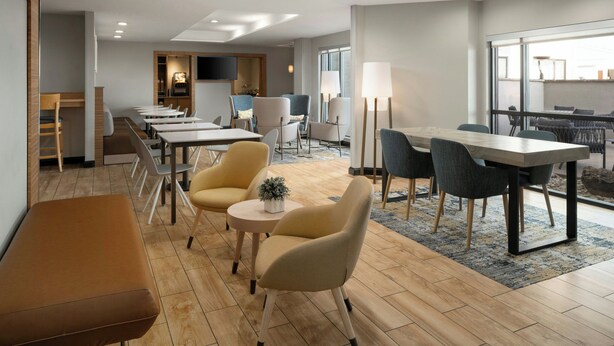 A modern hotel lobby with a long reception desk, multiple seating areas with tables and chairs, a flat-screen TV mounted on the wall, and a large window offering a view of the outside. - TownePlace Suites by Marriott Waco South (Waco)