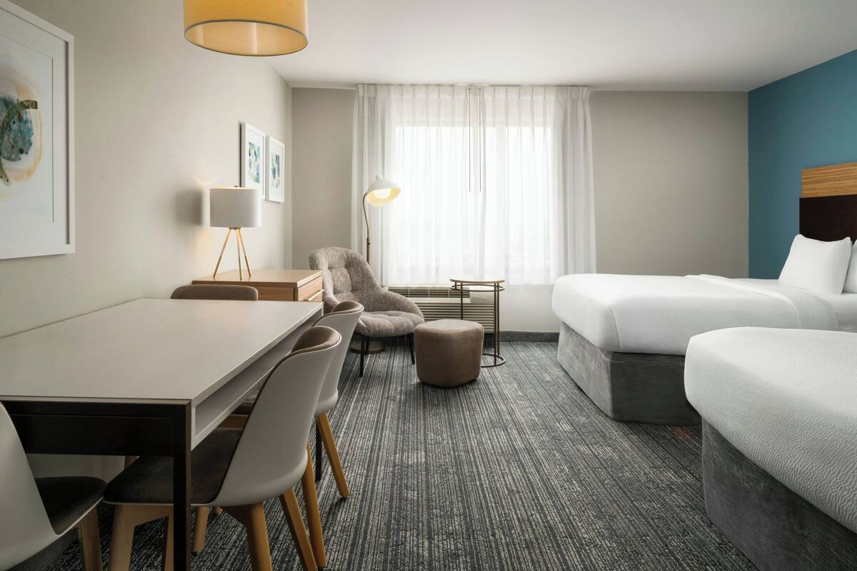 Habitación de hotel con cama, escritorio, sillas, lámpara y cuadros en las paredes.