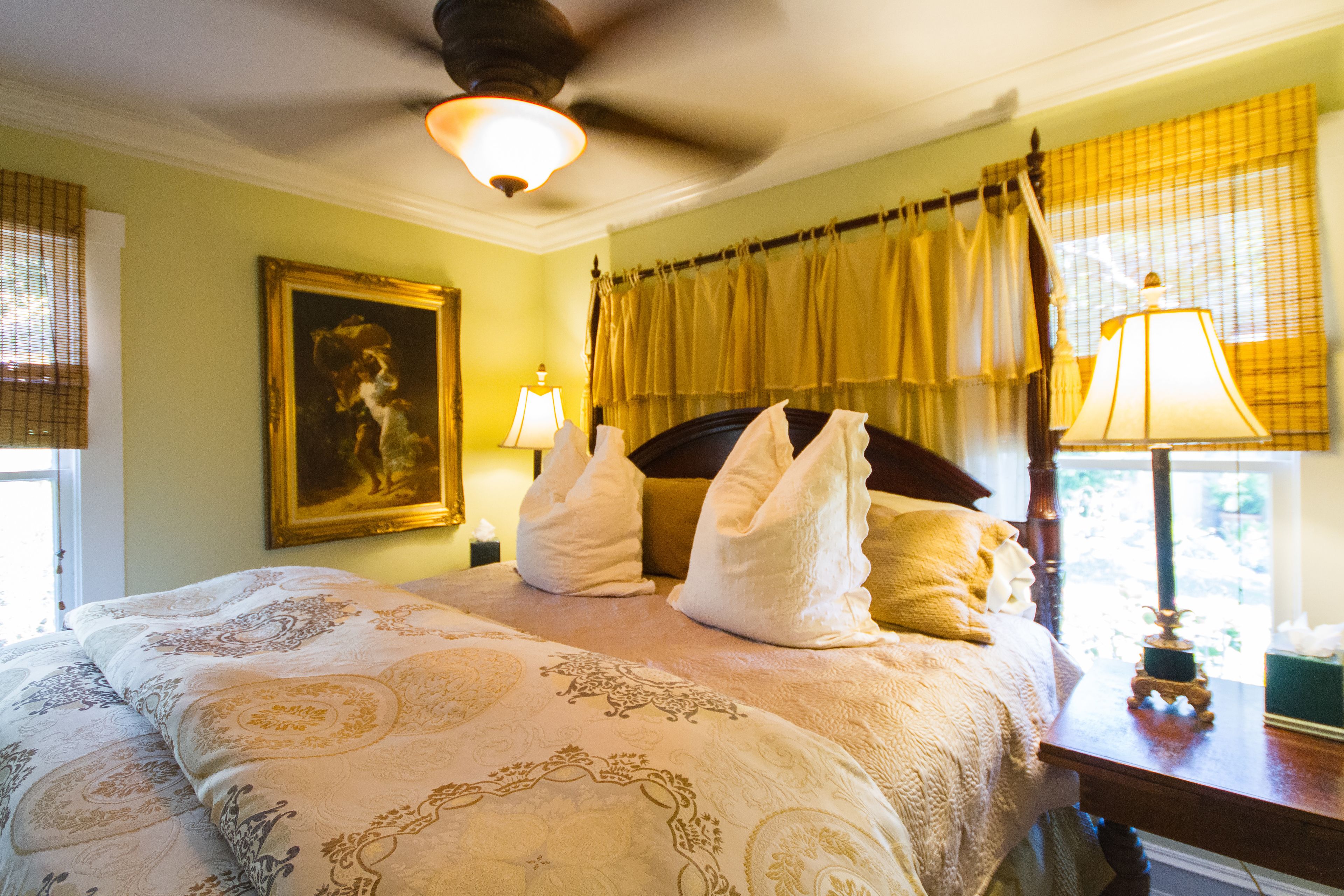 A hotel room with a large bed, a ceiling fan, a painting on the wall, and a window with curtains.