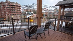 Un balcón con dos sillas, una terraza de madera y vistas a montañas nevadas y edificios.