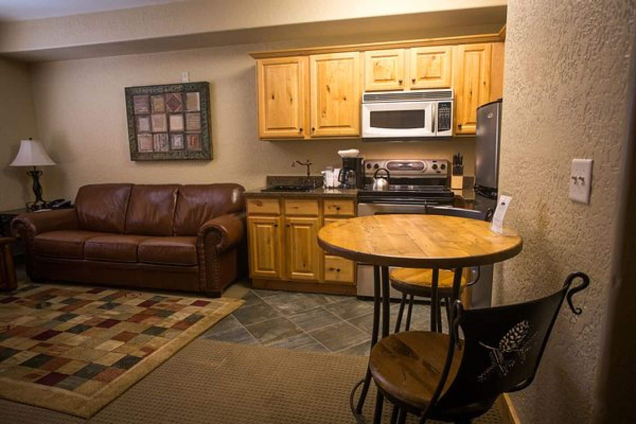 A compact living space with a kitchenette, a brown leather sofa, a round wooden table, and a black metal chair.