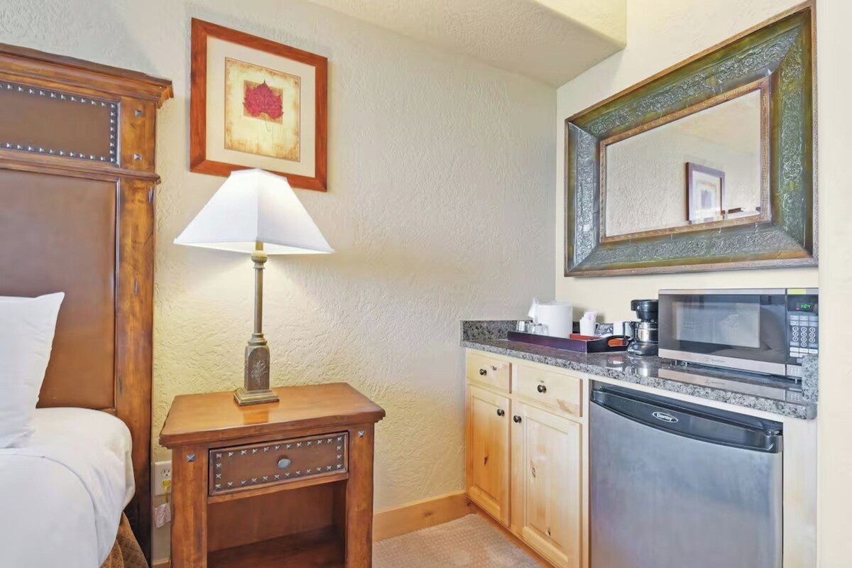 Quarto de hotel com cama, abajur, mesa pequena com luminária, micro-ondas e máquina de lavar louça.
