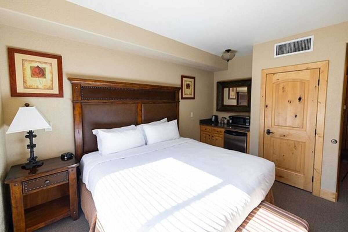 Habitación de hotel con una cama grande, una mesita de noche con lámpara, una puerta de madera y una pequeña cocina.