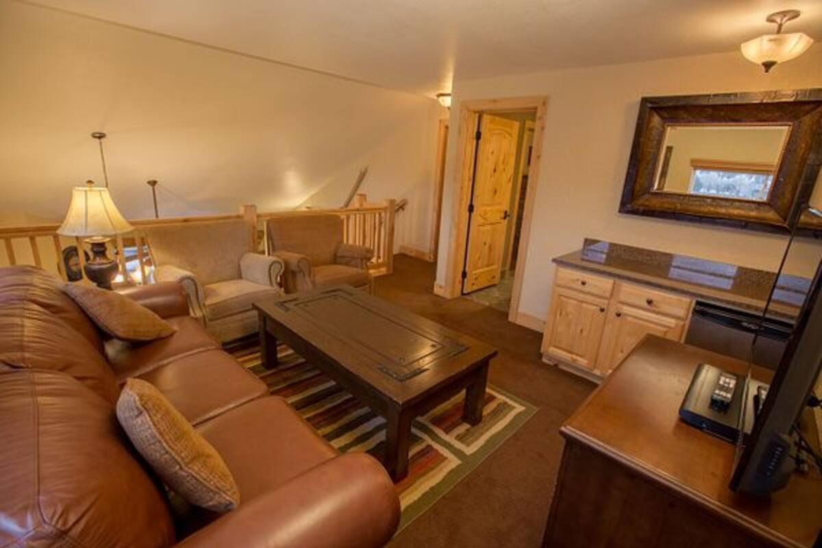 Una sala de estar con un sofá de cuero marrón, una mesa de centro de madera, una lámpara, un televisor y una zona de cocina con armarios y una encimera.