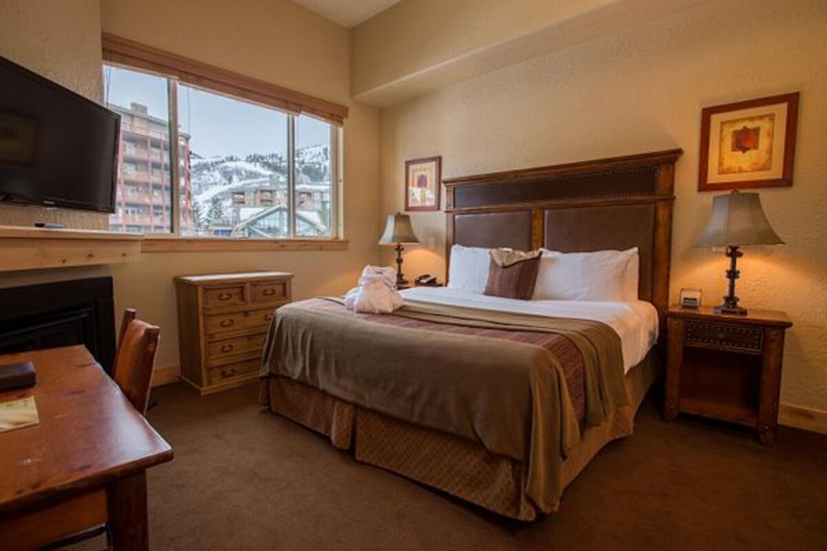 A hotel room with a bed, a desk, a television, a window with a view, and two bedside lamps.