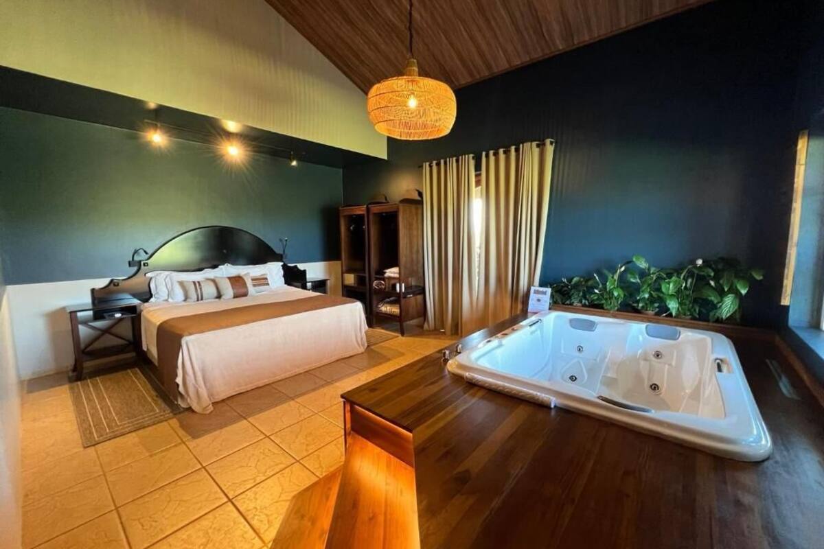 A hotel room with a bed, a wooden side table, a hot tub, and a hanging light fixture.