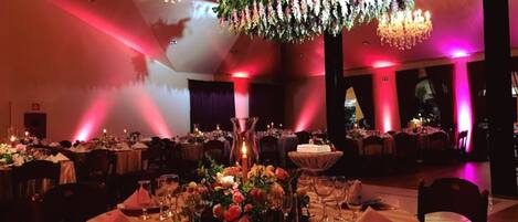 Een balzaal met ronde tafels gedekt voor een formeel evenement, met kroonluchters en bloemstukken als decoratie.
