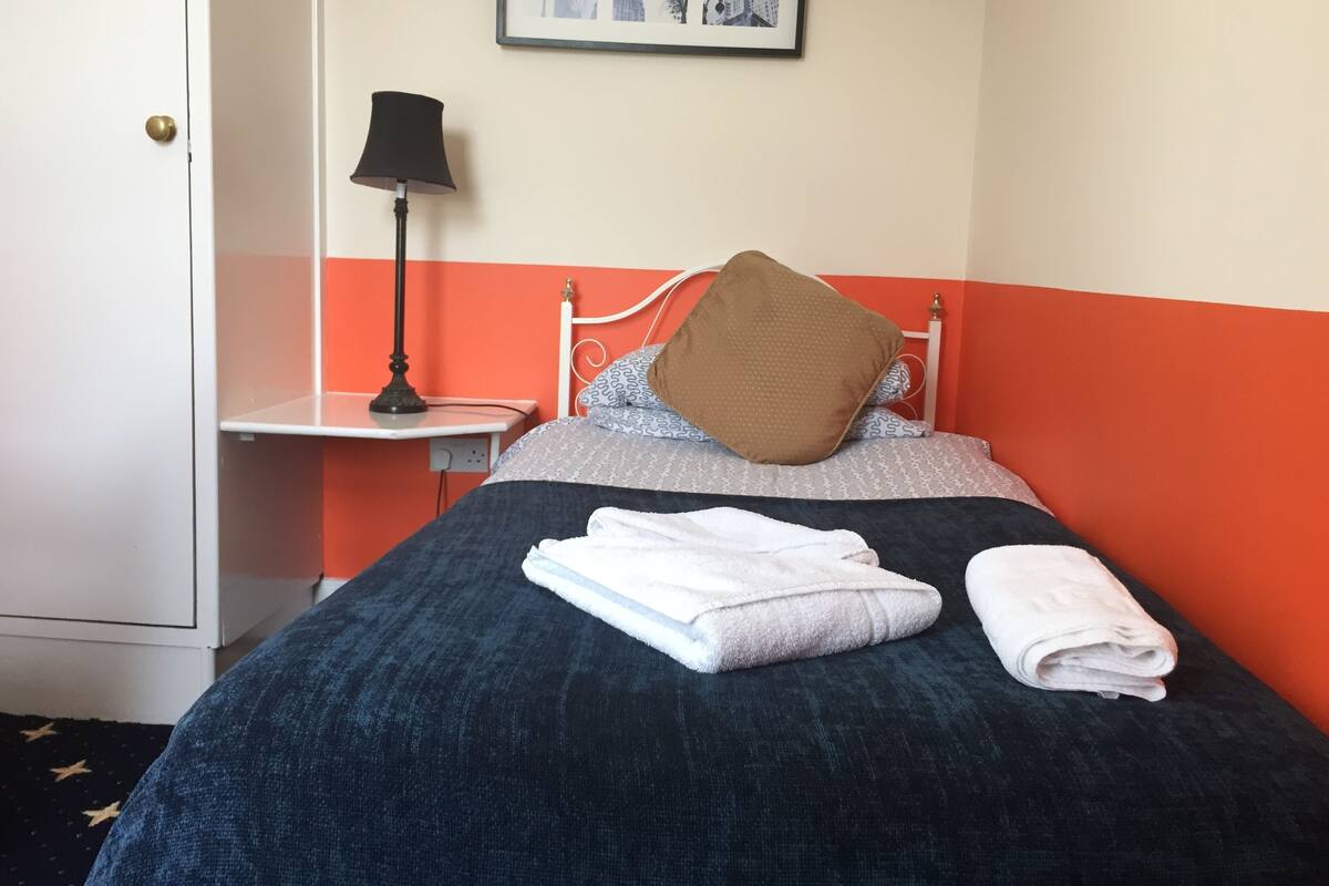 Une chambre d’hôtel avec un lit, une table de chevet avec une lampe, un tableau encadré au mur et une porte.