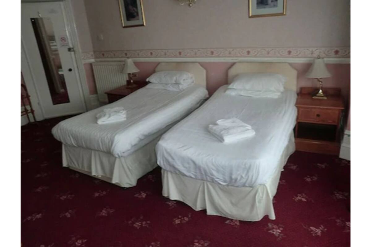 Une chambre d’hôtel avec deux lits simples, une moquette rouge, un radiateur et une table de chevet avec une lampe.