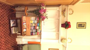 Een kamer met een open haard, een boekenkast, twee groene fauteuils en een bloemenvaas.