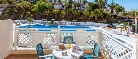 Uma área à beira da piscina com mesa e cadeiras, uma piscina e vista para palmeiras e edifícios ao fundo.