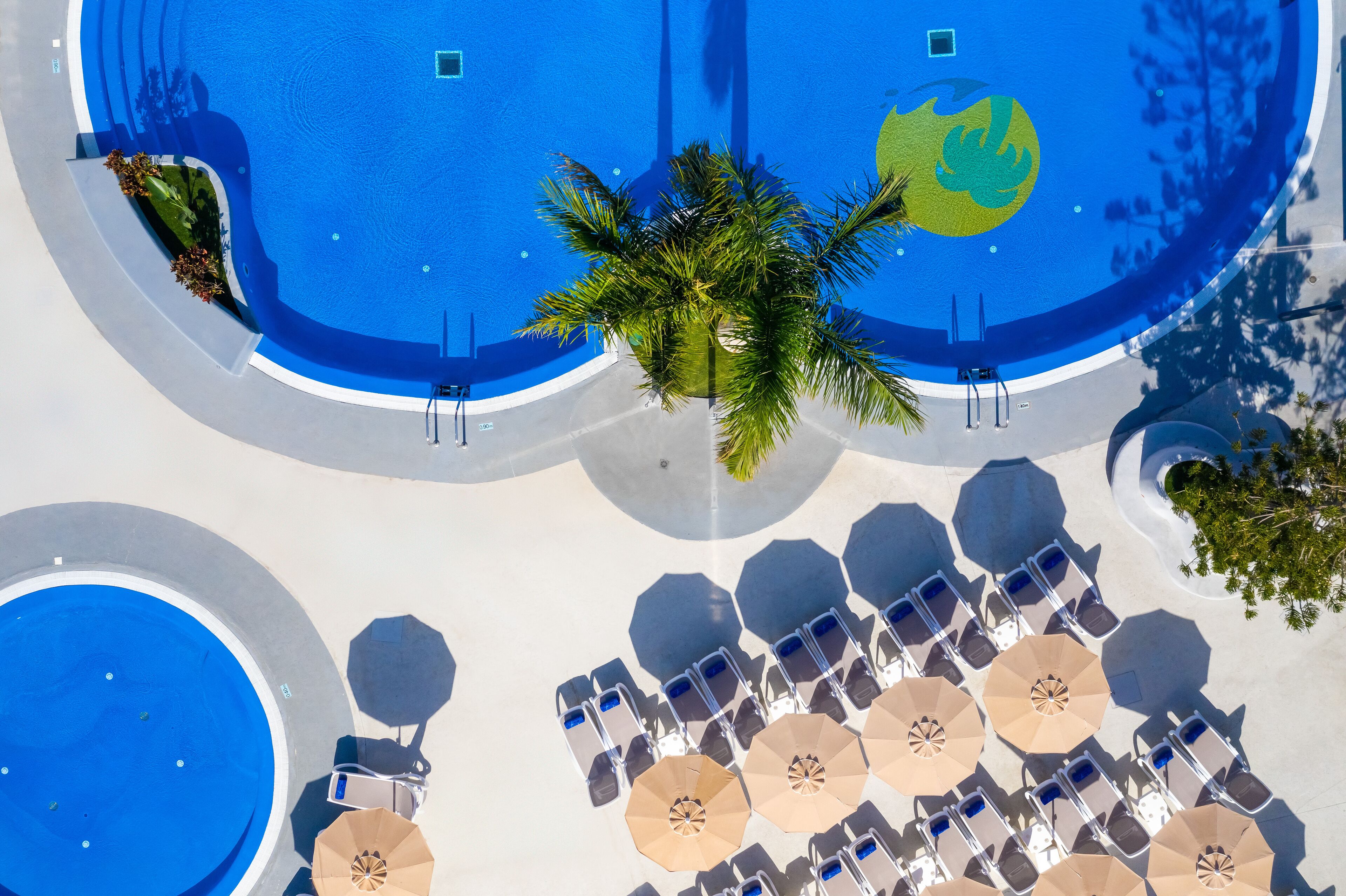 Vista aérea de un área de piscina en un resort, con múltiples sillas de descanso y sombrillas, rodeada de palmeras y una gran piscina azul.