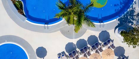 Vista aérea de un área de piscina en un resort, con múltiples sillas de descanso y sombrillas, rodeada de palmeras y una gran piscina azul.