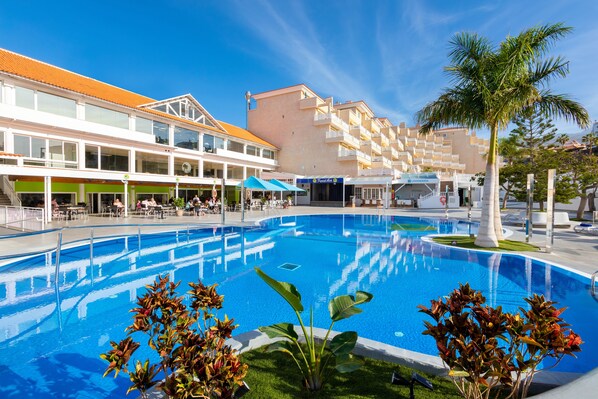 A swimming pool surrounded by buildings with balconies, a palm tree, and outdoor seating areas. - Hotel Tropical Park (Adeje)