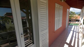 Un pasillo con puertas y contraventanas blancas, suelo de baldosas y vistas a edificios y vegetación en el exterior.