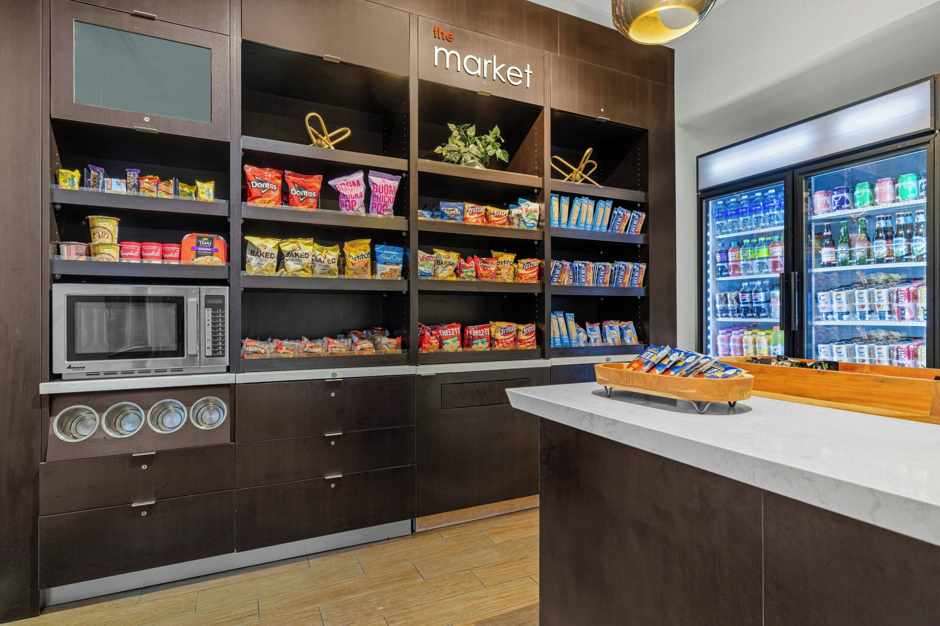 A kitchen area with a microwave, shelves stocked with snacks, and a refrigerator filled with beverages.