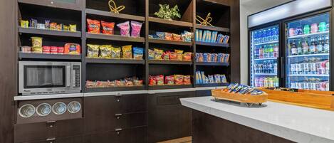 A kitchen area with a microwave, shelves stocked with snacks, and a refrigerator filled with beverages.