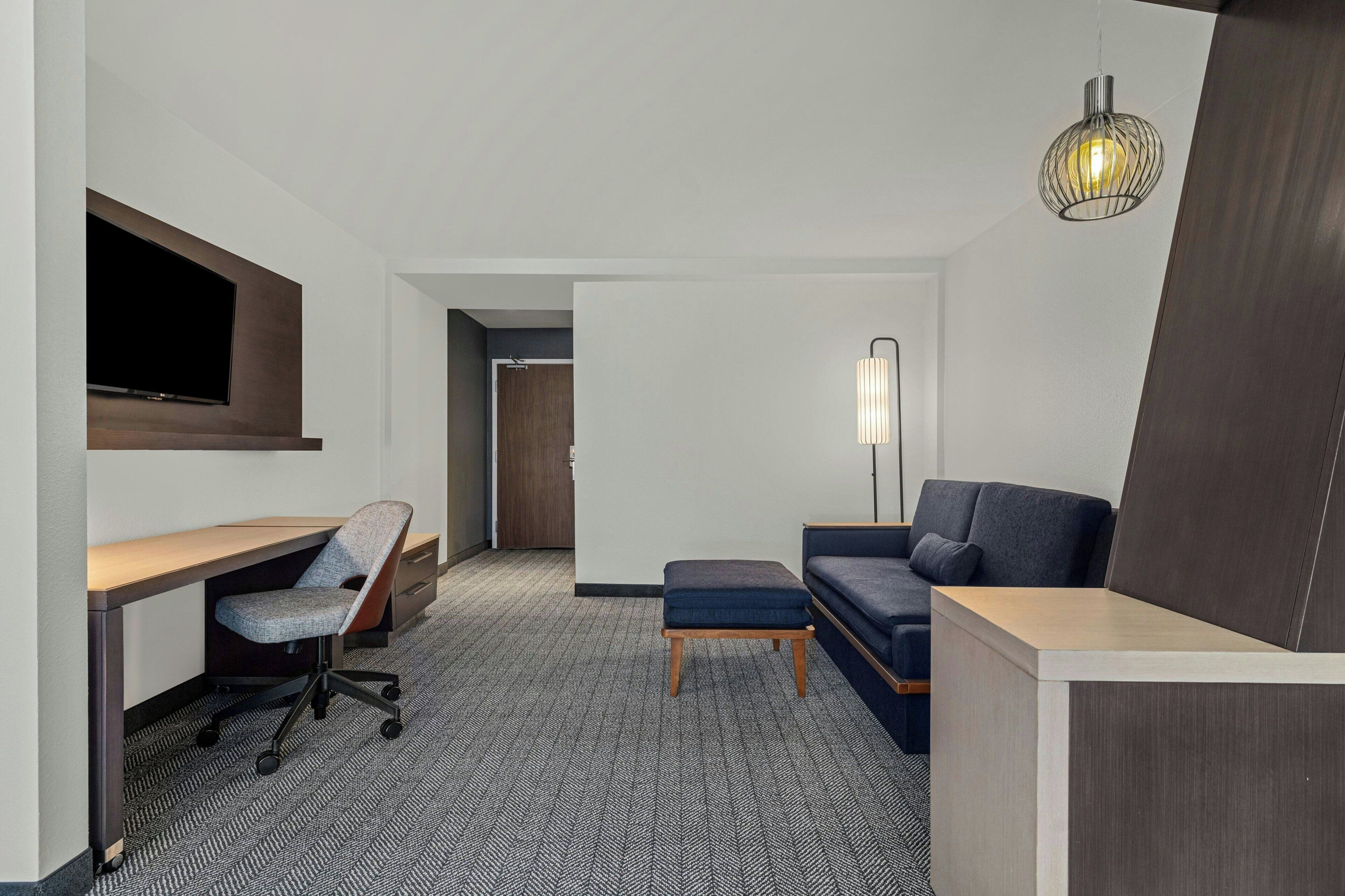 Une chambre d’hôtel équipée d’un bureau, d’une chaise, d’un canapé, d’un pouf et d’un téléviseur fixé au mur.