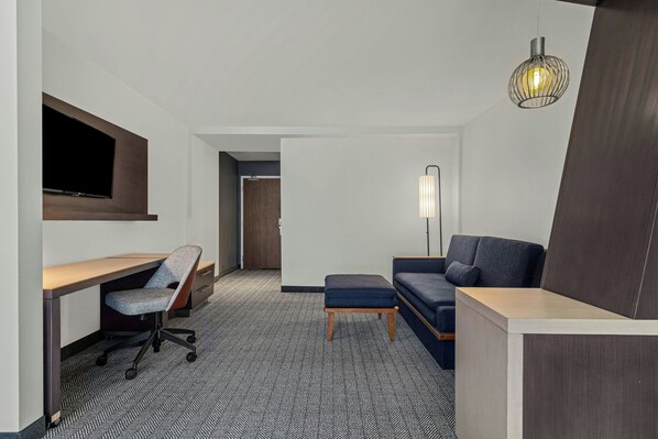 Habitación de hotel con escritorio, silla, sofá, reposapiés y televisor montado en la pared.