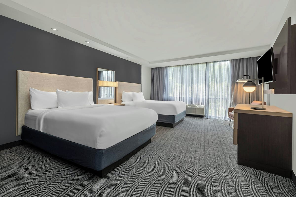 A hotel room with two beds, a desk, a TV, and a large window with curtains.