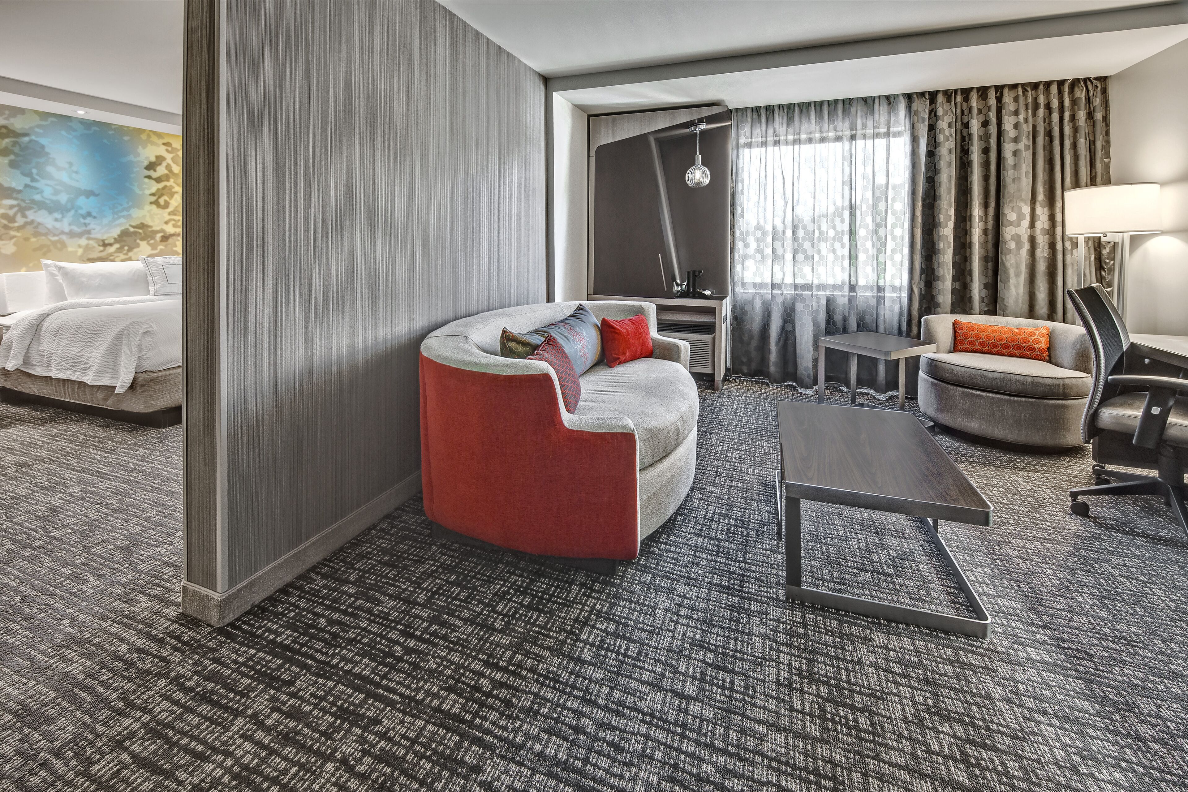 Une chambre d’hôtel moderne avec un tapis gris, un canapé gris agrémenté de coussins rouges et bleus, une petite table ronde, un fauteuil gris avec un coussin orange, une grande fenêtre avec des rideaux gris et un tableau accroché au mur.