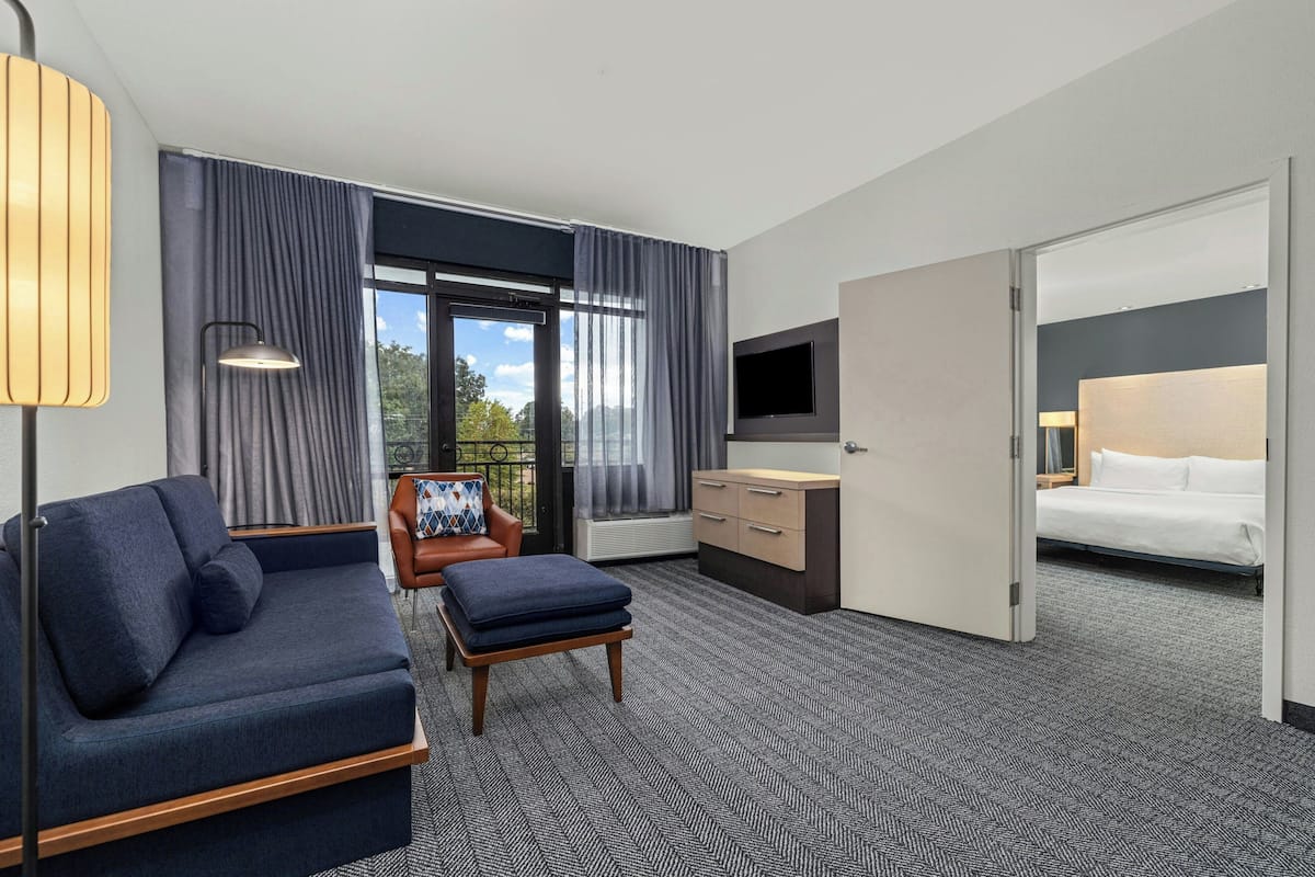 A modern hotel room with a sofa, armchair, and ottoman, a TV, a bed, and a large window with curtains.