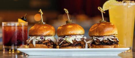 Een bord met drie pulled pork sandwiches met coleslaw, vergezeld van twee drankjes en een glas ijsthee.