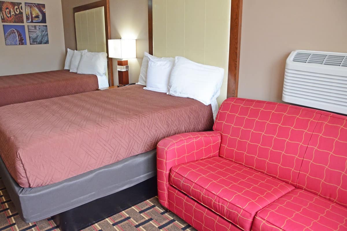 A hotel room with two beds, a red sofa, a lamp, and a wall-mounted air conditioner.