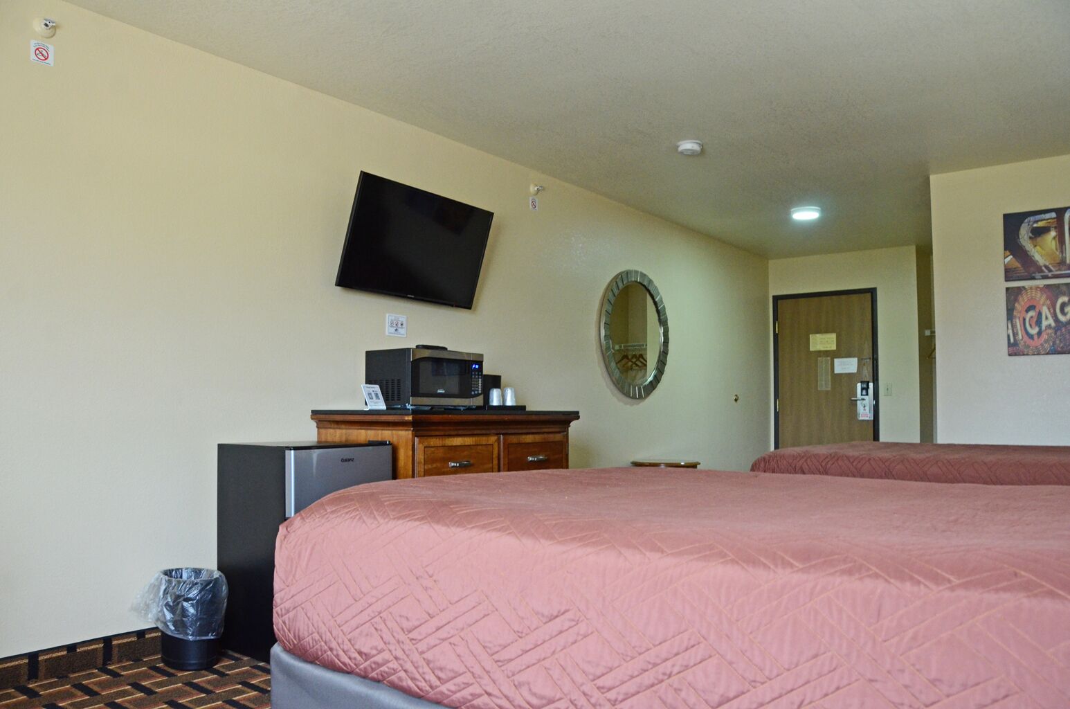 A hotel room with a bed, a television mounted on the wall, a microwave, a refrigerator, a trash can, and a door with a sign.