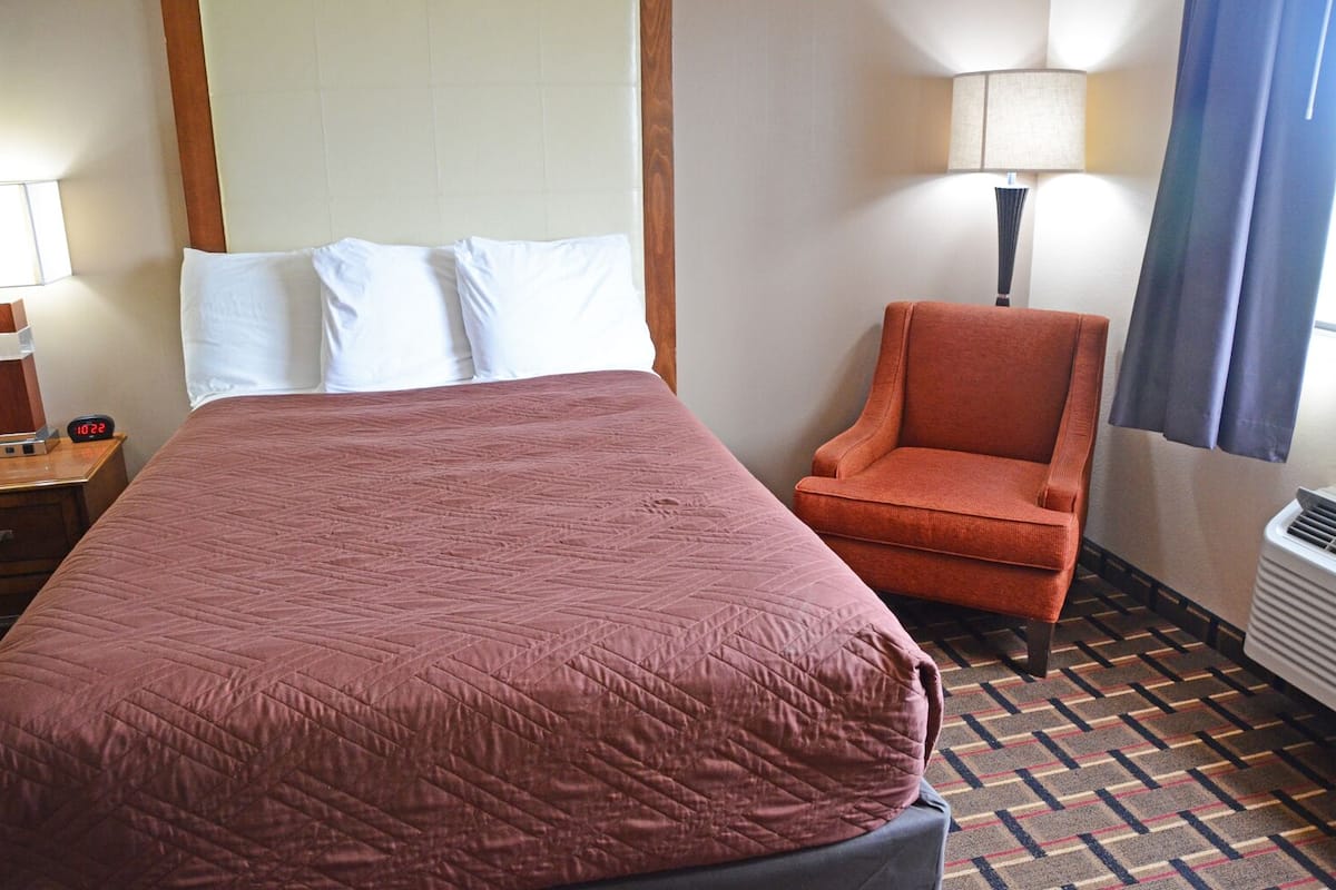 A hotel room with a large bed, a chair, a nightstand with a clock, and a lamp.