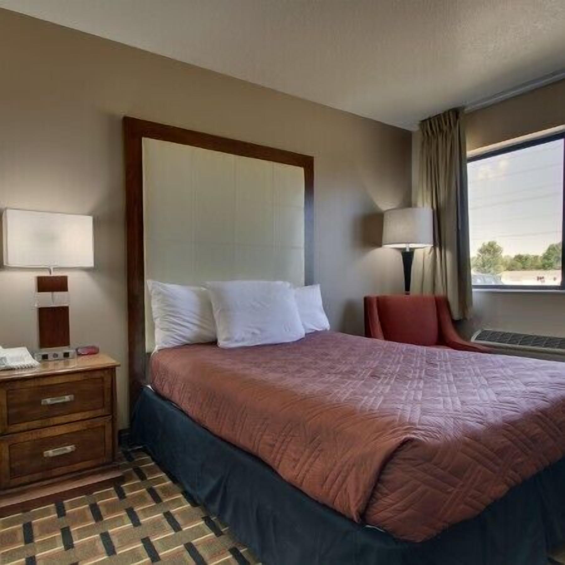 A hotel room with a large bed, a wooden nightstand with a lamp, a chair, and a window with a view of the outside.