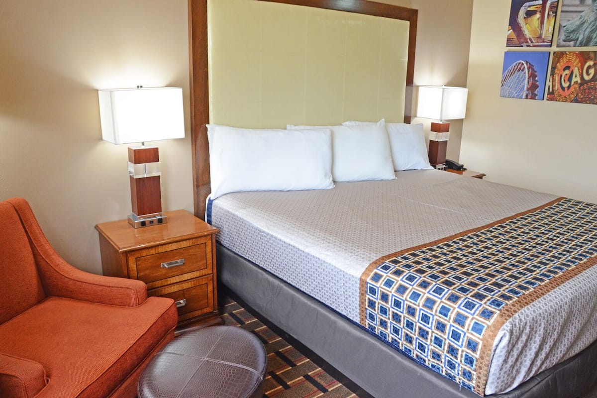 A hotel room with a bed, a nightstand, a chair, and a small stool.