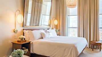 A modern hotel bedroom with a large bed, a round side table, a mirror, and a hanging light fixture.