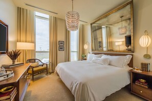 A hotel room with a large bed, a desk with a lamp, a chair, a bedside table with books, and a large mirror. - The Dewberry Charleston (Charleston)