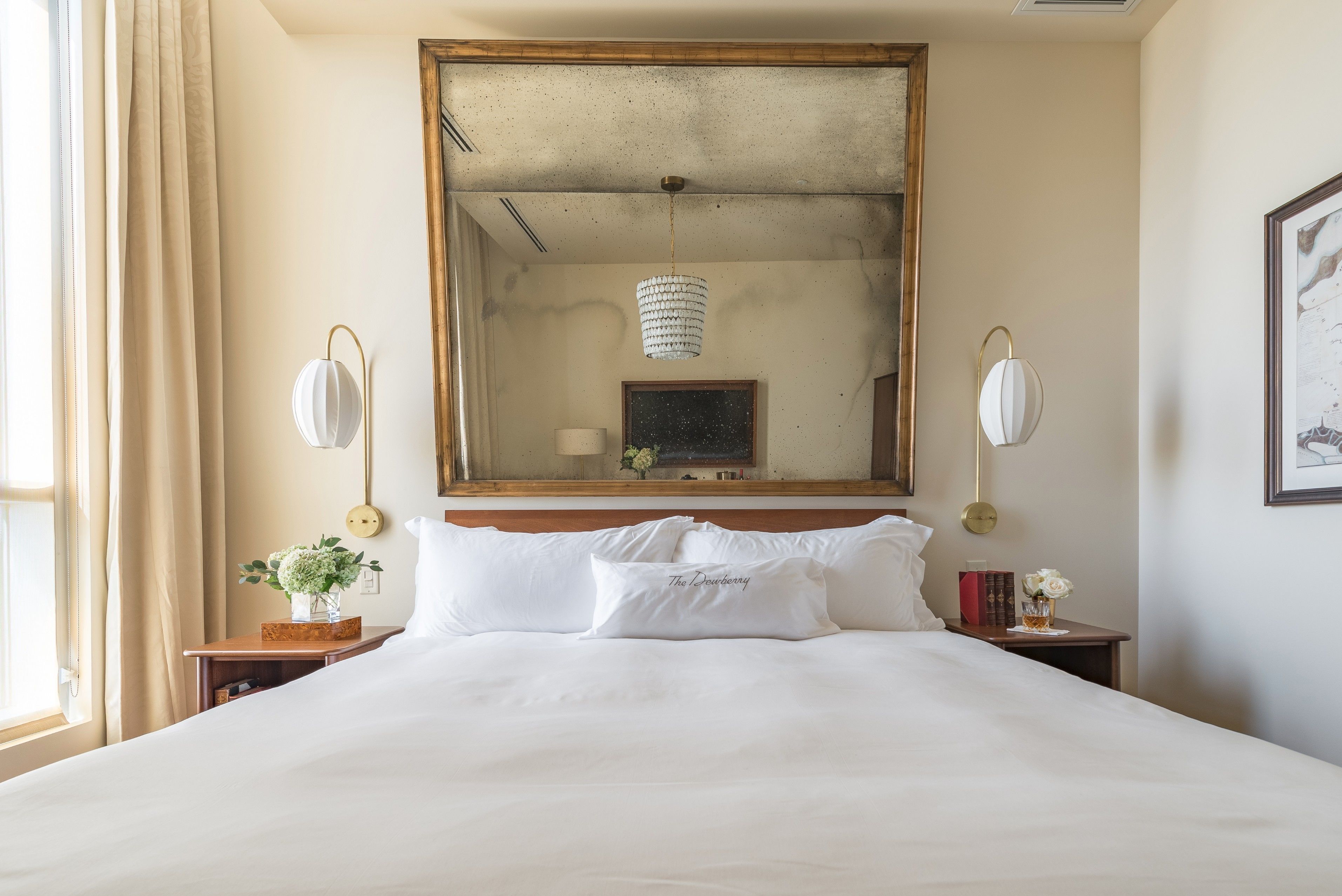 Una habitación de hotel con una cama grande, cabecera de madera, espejo, mesitas de noche con lámparas y una ventana con cortinas.