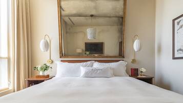 Una habitación de hotel con una cama grande, cabecera de madera, espejo, mesitas de noche con lámparas y una ventana con cortinas.